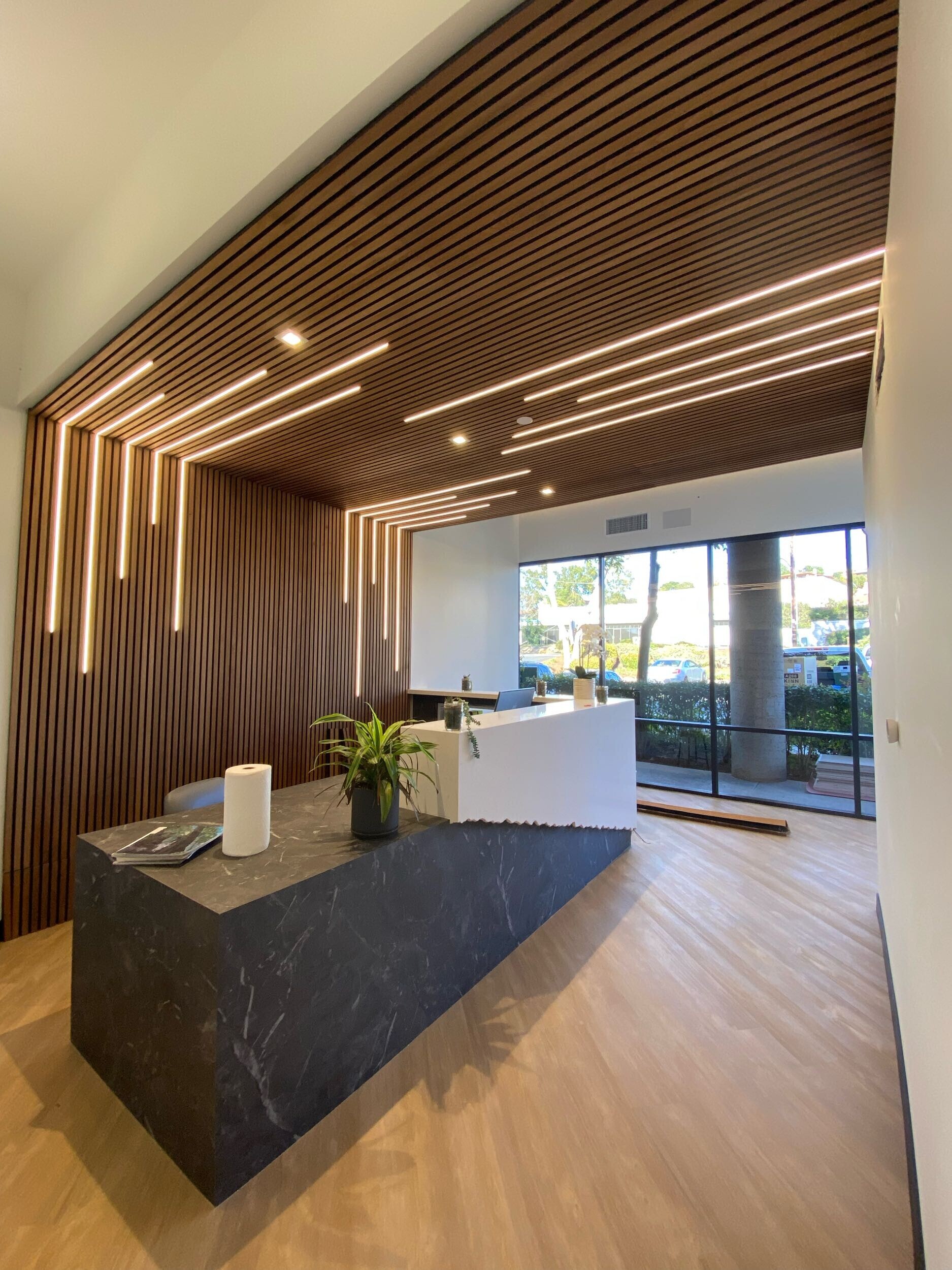 Modern office reception area with a dark marble desk, wooden slatted walls, and ceiling with linear lighting. open view to the outdoors on the left.